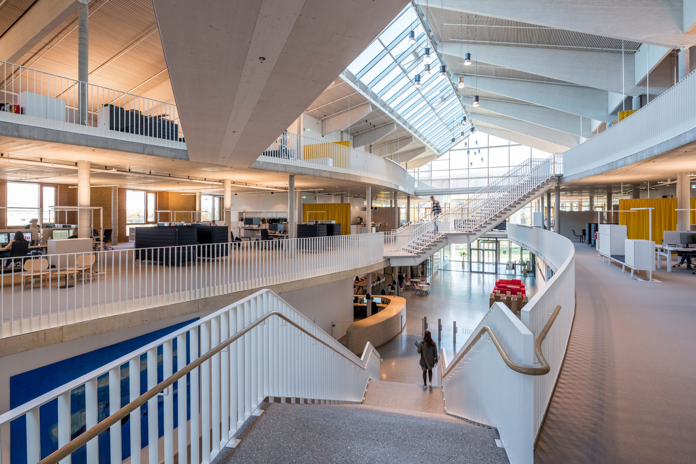 Blick in den Alnatura Campus in Darmstadt. Ein zweistöckiger Gebäudebereich, durch Treppen verbunden zeigt eine helle und offene Bürolandschaft und einige Arbeitsplätze.