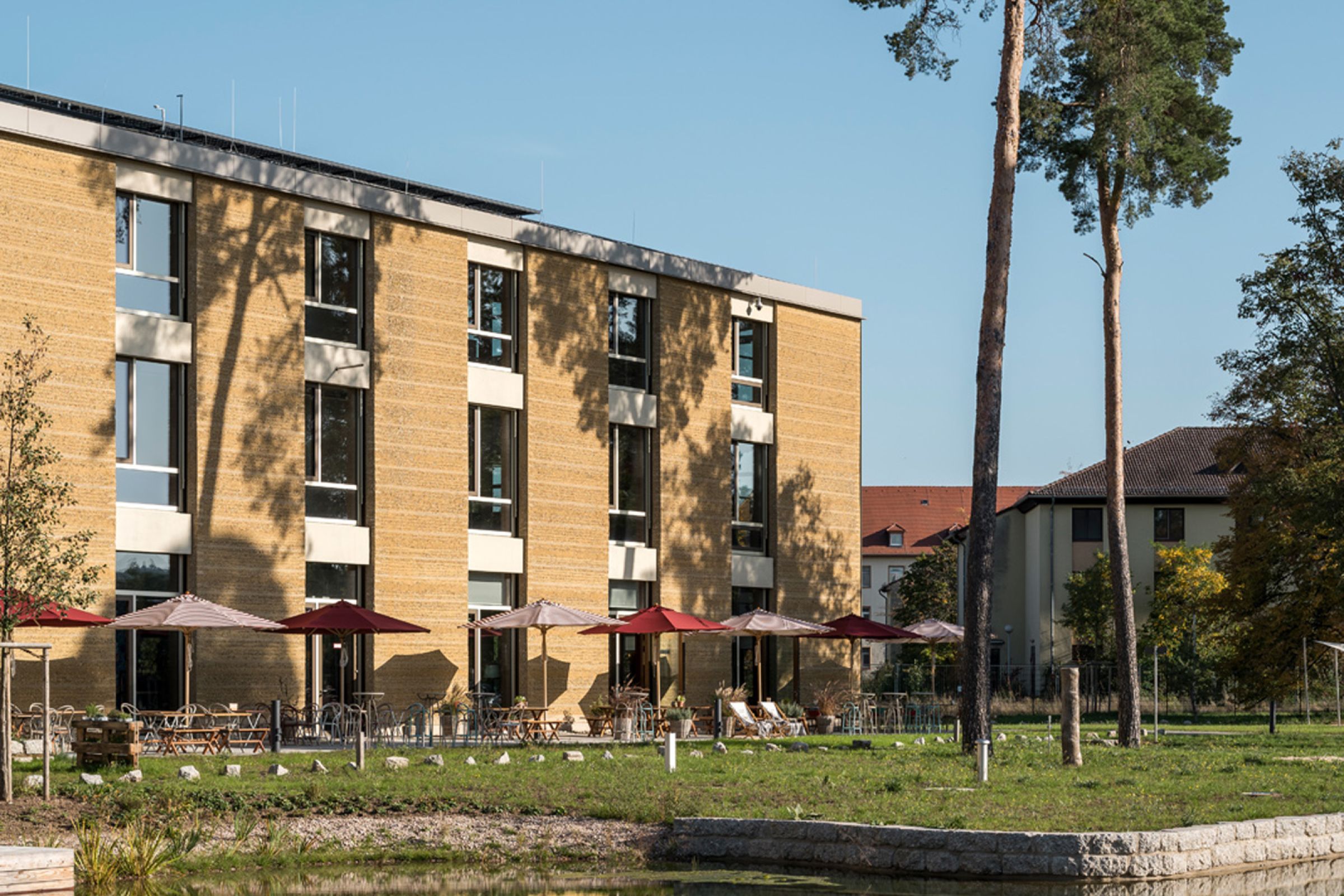 Außenansicht auf den Alnatura Campus in Darmstadt mit Sonnenschirmen sowie Sitzgelegenheiten vor dem Gebäude. Außerdem ist ein kleiner See vor dem Gebäude zu sehen.