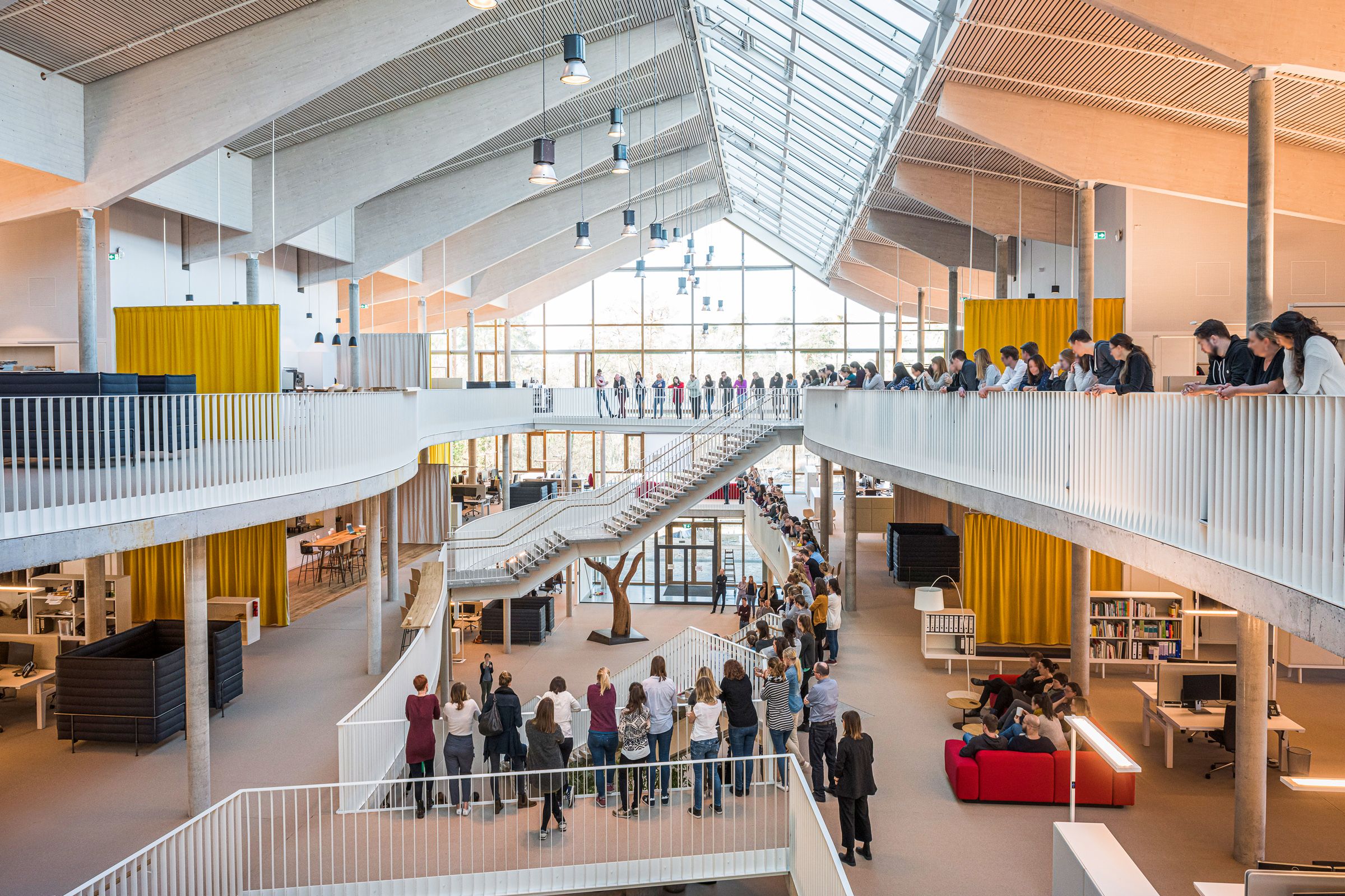 Sehr viele Mitarbeitende stehen auf verschiedenen Ebenen des Alnatura Campus in Darmstadt. Der Campus ist eine helle und offene Bürolandschaft, die durch Treppen verbunden ist.