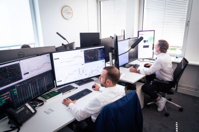 Zwei Mitarbeitende sitzen im Büro am Schreibtisch und schauen in große Bildschirme mit diversen Charts und Zahlen.